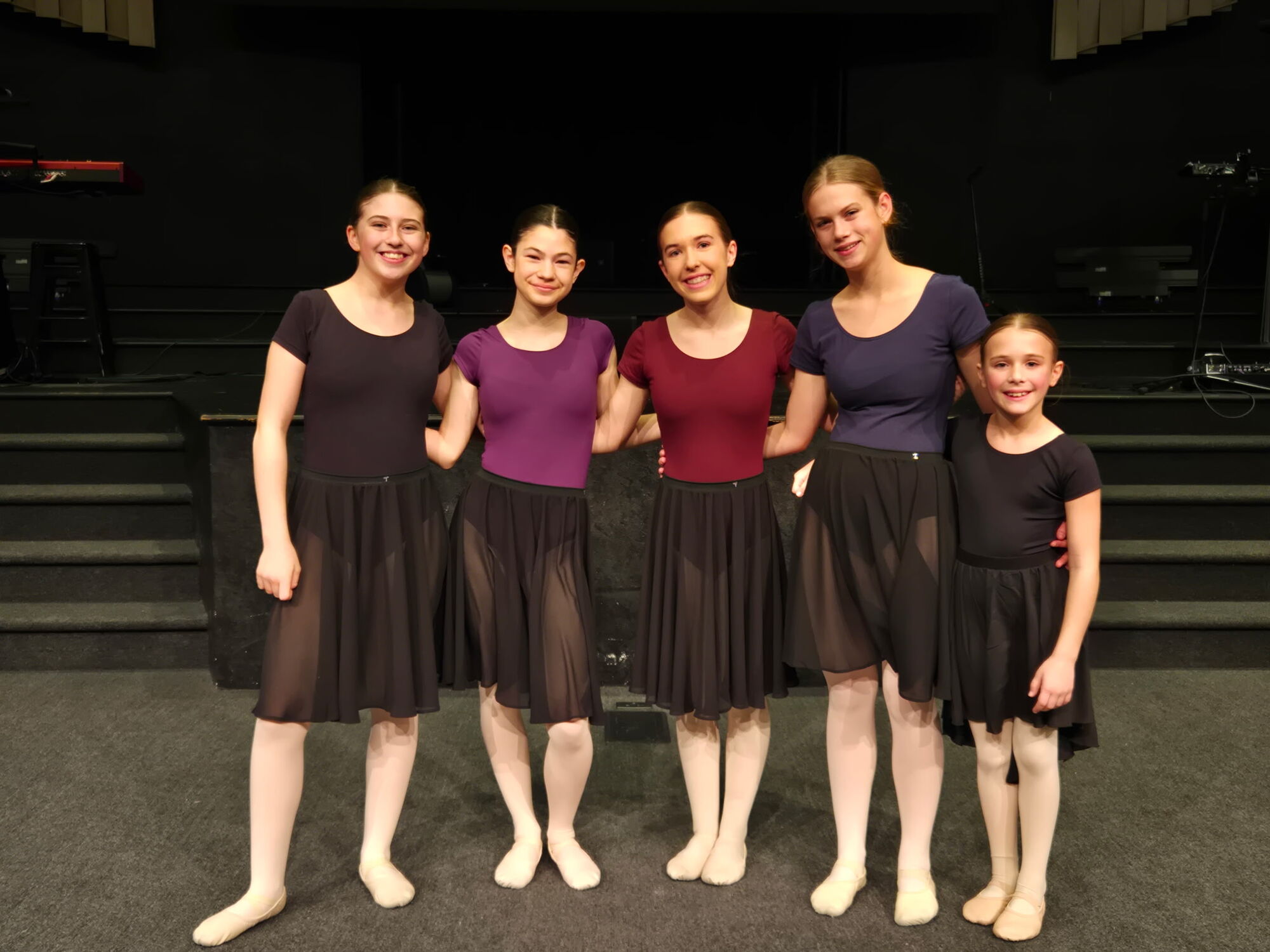 Five students in leotards and wrap skirts smiling arm-in-arm after class