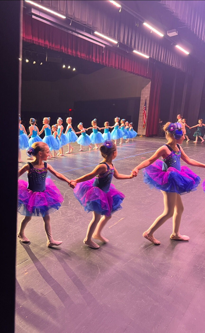 Young dancers in sparkling tutus hold hands on stage, seen from the wings
